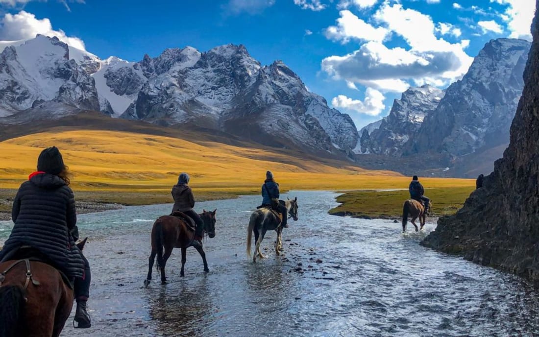 Travellers on horseback crossing a river toward snow-capped peaks